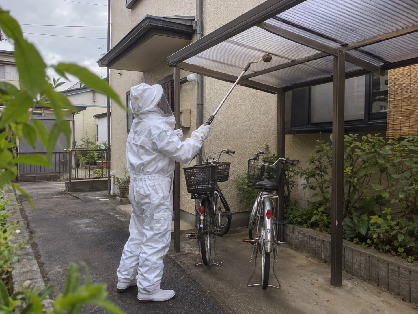 防護服の作業員が安定した地面から長い道具で自転車置き場の屋根下を処理する様子