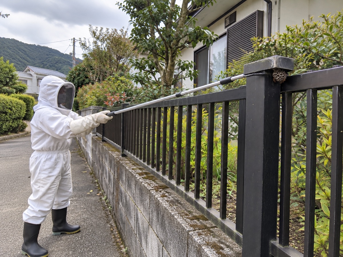 防護服の作業員がフェンスから距離を取り長い道具で巣を処理する様子